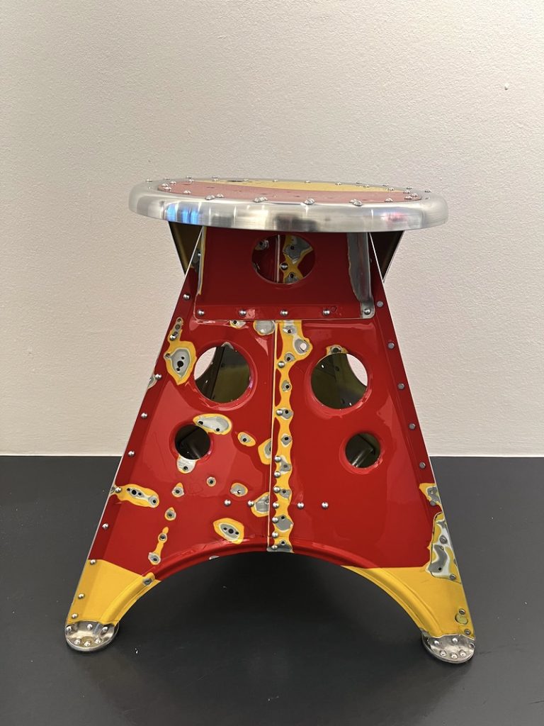 Hocker gefertigt aus dem Schrott alter Hubschrauber und Flugzeuge. Bei Wartungsarbeiten anfallende Bleche älterer Modelle lassen sich teilweise zu leichten Hockern recyceln, deren unterschiedliche Farben, Beschriftungsspuren und Nietlöcher noch auf die ursprüngliche Verwendung verweisen. So entsteht als Unikate in Handarbeit ein leichtes (1,4kg) Möbel mit hoher Belastbarkeit (250kg). Entstanden ist ein schlichtes Möbel, das trotz filigraner Konstruktion extrem stabil ist. Punktuelle, lineare und flächige Elemente, resultierend aus dem Formen zweidimensionalen Bleches zu einem dreidimensionalen Körper, führen zu einem Objekt von spezifisch konstruktiver Ästhetik mit interessanten Ein- und Durchblicken. (Nominiert für den Staatspreis NRW, 2013)